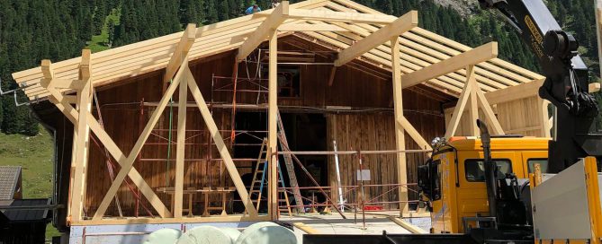 Holzbauarbeiten an Almhütte mit offenem Dachstuhl, Kran, Bauholz und grüner Almlandschaft