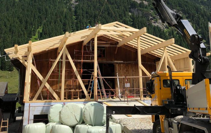 Holzbauarbeiten an Almhütte mit offenem Dachstuhl, Kran, Bauholz und grüner Almlandschaft
